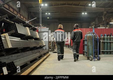 Rear view of two young interracial male workers in uniform discussing quality of new equipment while moving along aisle in warehouse Stock Photo