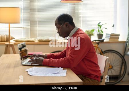 African businessman sitting at table with documents and doing online work using laptop in the room Stock Photo