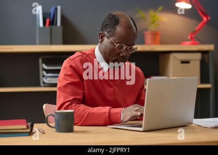 African mature businessman in eyeglasses concentrating on his online work using laptop while sitting at office desk with cup of coffee Stock Photo