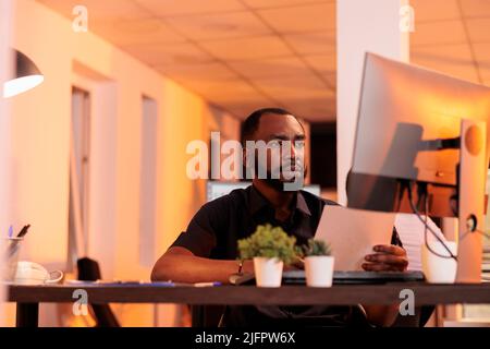 Executive manager analyzing paperwork report and taking notes to create research presentation in office with big windows. Working on project data with documents and computer during sunset. Stock Photo