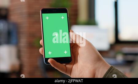 Male freelancer holding modern smartphone with greenscreen display, looking at blank mockup template. Working with isolated chroma key copyspace on mobile phone background in office. Stock Photo