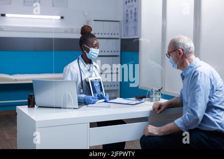 African american radiology specialist explaining x-ray scan image to senior patient while wearing virus protection facemasks. Expert radiologist showing MRI scan image to elderly man. Stock Photo
