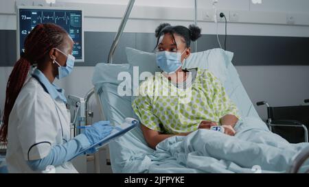 Medical specialist doing healthcare checkup with sick woman in bed during coronavirus pandemic. Physician taking notes and having conversation with patient, wearing face masks. Stock Photo