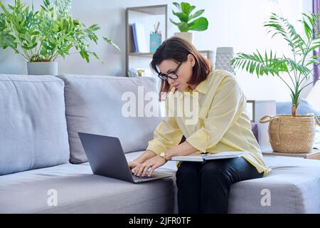 Mature business woman working on couch, using laptop making notes in notebook Stock Photo