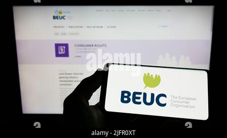 Person holding mobile phone with logo of Bureau Europeen des Unions de Consommateurs (BEUC) on screen in front of webpage. Focus on phone display. Stock Photo