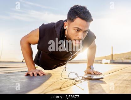 Push if you want to achieve your goals. Shot of a sporty young man wearing earphones while doing push-ups, on a rooftop. Stock Photo