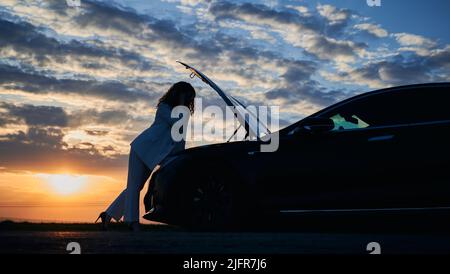 Silhouette of businesswoman looking under the hood of car while standing in field with beautiful evening sky on background. Young woman in stylish suit checking car engine under the hood during sunset Stock Photo