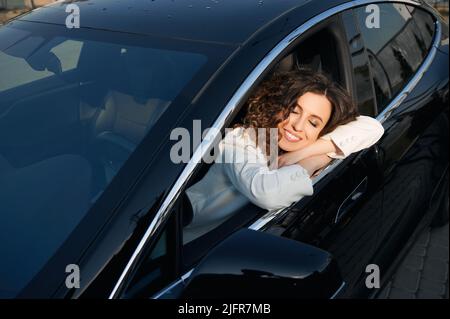 Beautiful woman sitting on driver seat in electric car and smiling while using vehicle autopilot system during her sleep. Cheerful businesswoman sleeping in electro car with driver-assistance system. Stock Photo