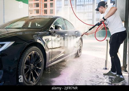Casual dressed professional serviceman cleaning black car during work time in outdoors carwash. Caucasian male in cap using self-service during daytime in city for washing own automobile vehicle Stock Photo