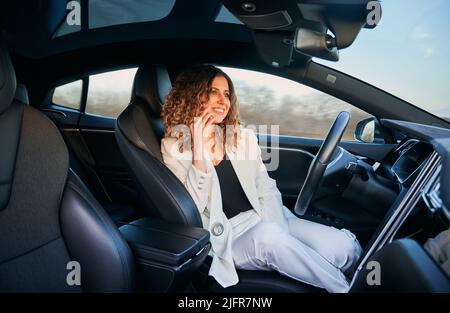 Young woman having phone conversation and smiling while riding automotive self driving in electric car. Joyful businesswoman talking on mobile phone, using autopilot trip with hands free mode. Stock Photo