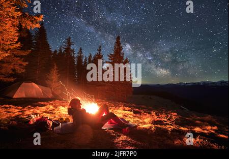 Female hiker lying at karemat near bonfire admire beauty of stunning starry sky during night in campsite, backpacker visiting mountains with tent near spruce forest in National Park Stock Photo