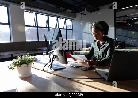 African american young businesswoman examining documents while sitting at computer desk in office Stock Photo