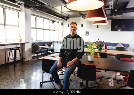 Portrait of smiling young caucasian businessman sitting on desk in office Stock Photo