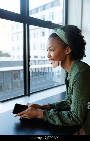 Smiling thoughtful african american businesswoman with smart phone looking through window in office Stock Photo