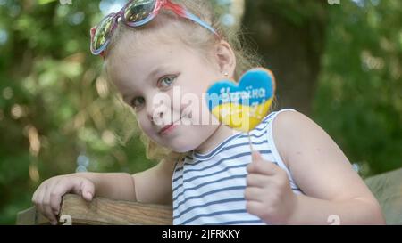 Little blonde girl holds gingerbread in Ukrainian national colors in her hand, it says 'Ukraine is me'. Close-up portrait of girl sitting on park benc Stock Photo