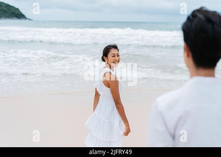 Happy smiling Asian 30s woman turning around and being called by man from back on the sandy beach in summer. Couple wearing a white shirt and dress - Romantic relationship and travel together concept Stock Photo
