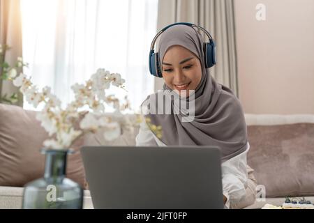Asia muslim lady wear headphone watch webinar listen online course communicate by conference video call at night home office. Remotely working from Stock Photo