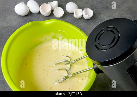 Beaten eggs with sugar in a green bowl with a beater immersed in a mixer and eggshells from chicken eggs on a dark concrete kitchen table. Making bize Stock Photo