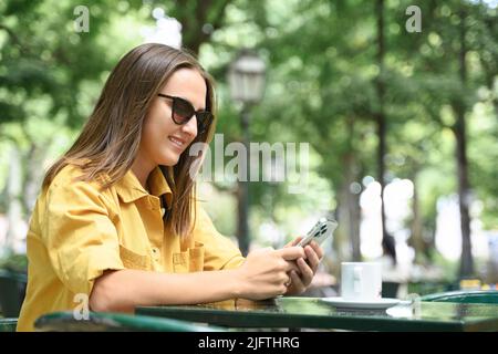 Side view at smiling young woman using smartphone outdoors, texting online during coffee break in summer cafe, positive smiling lady in sunglasses and yellow casual shirt spends time in social network Stock Photo