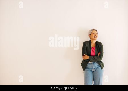 Young businesswoman laughing cheerfully while standing with her arms crossed. Happy young businesswoman standing against a wall background in a modern Stock Photo