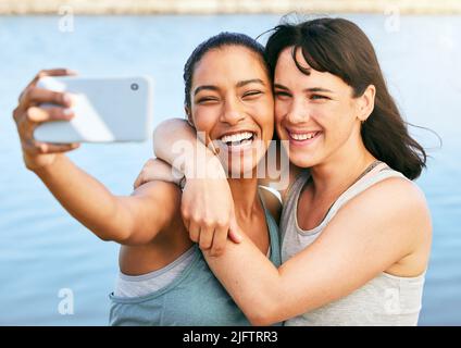 Two female friends taking selfies after a workout in nature and standing in front of a lake using a smartphone. Young Latino female using her phone to Stock Photo