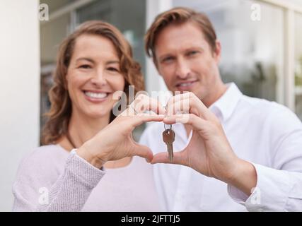 Closeup hands of happy married couple holding up new house keys with a heart shape hand gesture. Man and woman holding keys to new house or apartment. Stock Photo