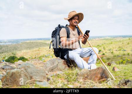 Middle aged with hatusing mobile phone while sitting on top of hill during trekking - concept of internet connection, technology and social media Stock Photo