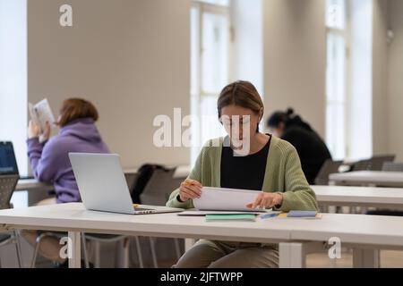 Hard-working female mature student reading architectural drawings while studying in public library Stock Photo