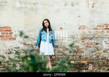 A teenage girl with freckles and long dark stright hair outside leaning against a brick wall and looking at the camera Stock Photo