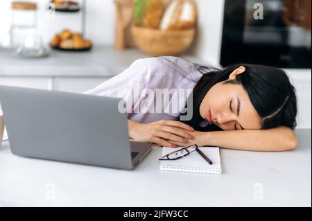 Sleepy chinese ethnicity young woman, fall asleep while seated at workplace near laptop at home in the kitchen, tired of boring job, overworked, experiencing chronic sleep deprivation, need rest Stock Photo