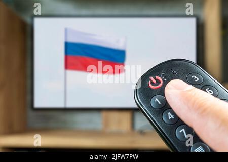 Remote control in a man's hand on the background of a TV and a Russian flag. The concept of disconnecting Russian channels in Europe and the USA. Bloc Stock Photo