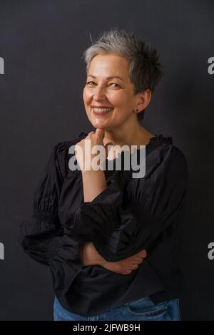 Mature businesswoman with grey hair smiling posing leaned chin on folded hands isolated on black background. Graying elegant woman with short hair. Image of successful mature woman. Aged beauty. Stock Photo