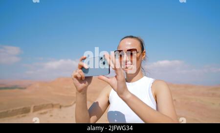 A young pretty woman who shoots a beautiful landscape. Action. A woman in sunglasses standing in the mountains makes a video on a new Phone and the bl Stock Photo
