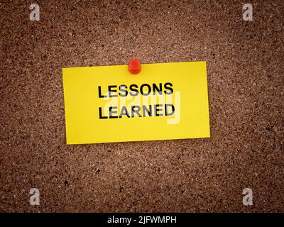 A paper note with the words Lessons Learned written on it pinned to a cork board. Close up. Stock Photo