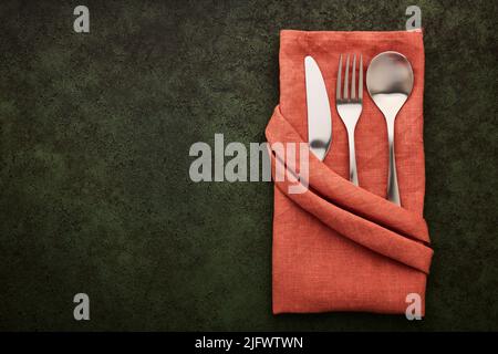 Terracotta-colored linen napkin folded in the shape of a pocket with cutlery inside, dark green background with copy space Stock Photo