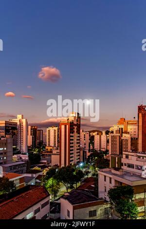 District of the city of Belo Horizonte in Minas Gerais with its buildings lit up by the sunset and with the full moon rising in the background Stock Photo