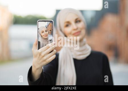 Focus on smartphone screen. Smiling cute young arabian islamic woman in hijab doing selfie on her smart phone for social media in city, outdoor, empty copy space. Stock Photo