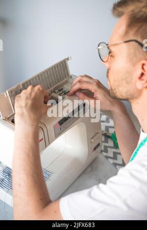A blond man with glasses changes a bobbin in a sewing machine. A tailor with a measuring tape on his neck in a white t-shirt.Concept of small business Stock Photo