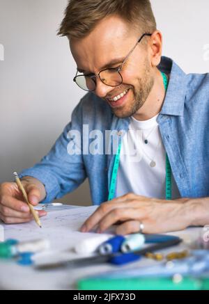 A DIY designer draws a sketch for a new costume project. A seamster with glasses is working at the table. A smiling man in a blue shirt makes a sketch Stock Photo