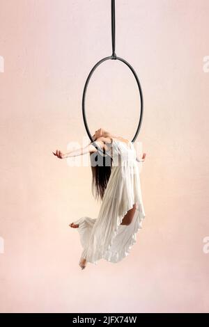 Full body of female aerialist in dress performing cross pose on aerial hoop during rehearsal against light background in studio Stock Photo