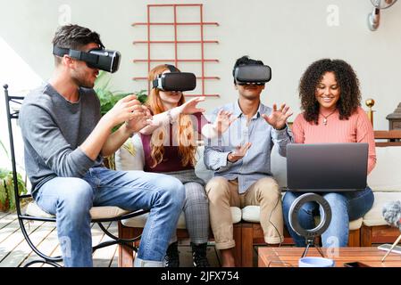 Multiracial men and women in casual clothes with VR helmets interacting with cyberspace while sitting near girlfriend browsing game on laptop on sunny Stock Photo