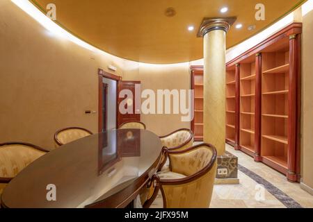 Professional office with round wooden table with wooden and yellow fabric armchairs and wooden bookshelf on oval wall painted in solid color Stock Photo