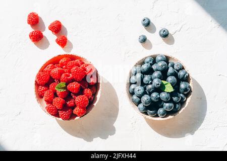 Top view fresh blueberries and raspberries in ceramic bowls on white textured background in sunlight. Healthy eating, vegan food flat lay. Stock Photo