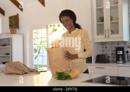 African american young woman removing fresh vegetables from shopping bag in kitchen at home Stock Photo