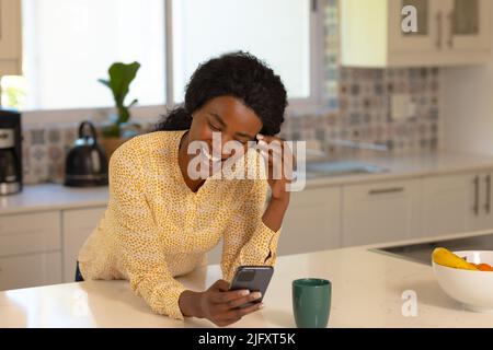 Cheerful young african american woman using smart phone while leaning on kitchen island Stock Photo