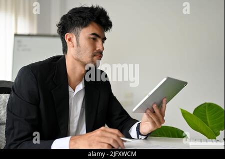 Smart and professional Asian businessman or male manager in formal suit sits at his desk, using digital tablet touchpad. Stock Photo