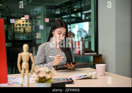 Successful and confident Asian female graphic designer manager using digital tablet to design her graphic artwork at her office desk. Stock Photo
