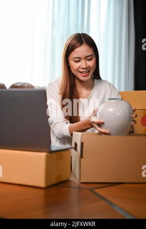 Charming millennial Asian woman online shop owner packing her goods into a cardboard box. Pottery online selling concept. Stock Photo