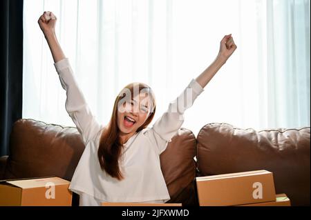 Cheerful and successful young Asian woman excited with her sales orders, hands up raised arms to cerebrating success. Stock Photo
