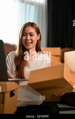 Portrait of a beautiful millennial asian female SME business owner updating her stock and shipping status on laptop computer while preparing a shippin Stock Photo